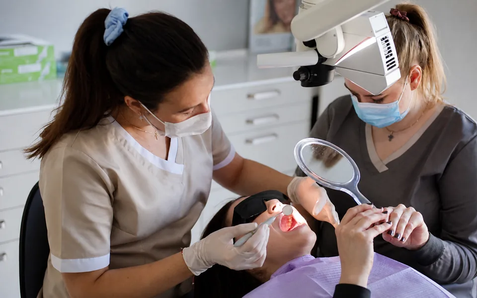 A dentist examining a patient's mouth while an assistant holds a mirror